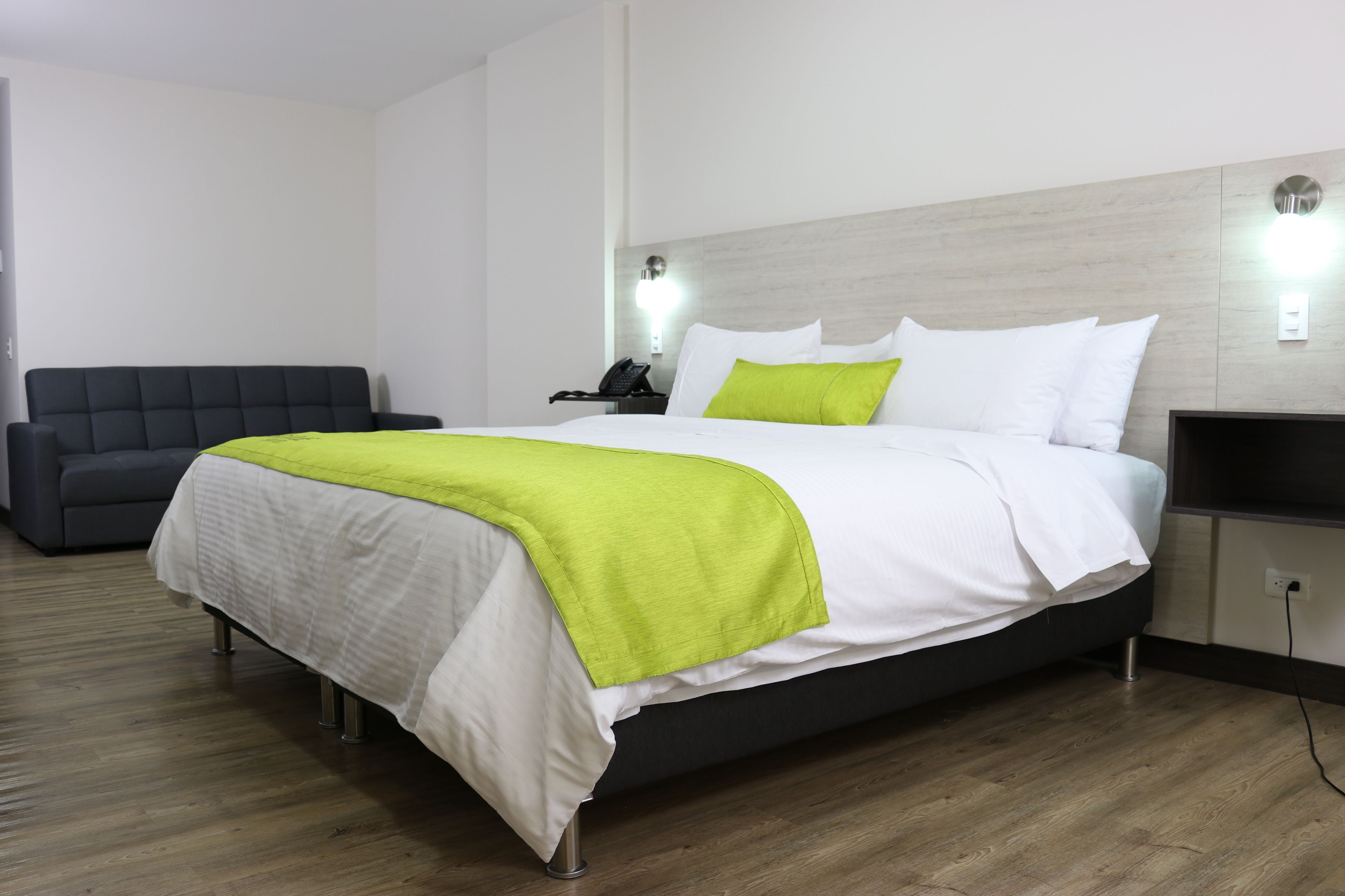Una habitación de hotel moderna con una cama, una mesita de noche, una lámpara y un sofá gris.