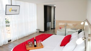 Habitación de hotel con cama, bandeja con una botella y vasos, almohada roja y vistas al exterior a través de cortinas traslúcidas.