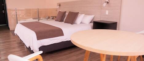 A modern hotel room with a large bed, bedside tables, a round wooden table, and a white chair.