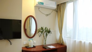 A hotel room with a flat-screen TV mounted on the wall, a wooden desk with a mirror, a potted plant, and a window with sheer curtains.