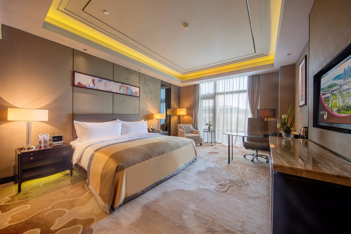 Ein Hotelzimmer mit einem großen Bett, Nachttischlampen, einem Schreibtisch, einem Stuhl und einem an der Wand befestigten Fernseher.