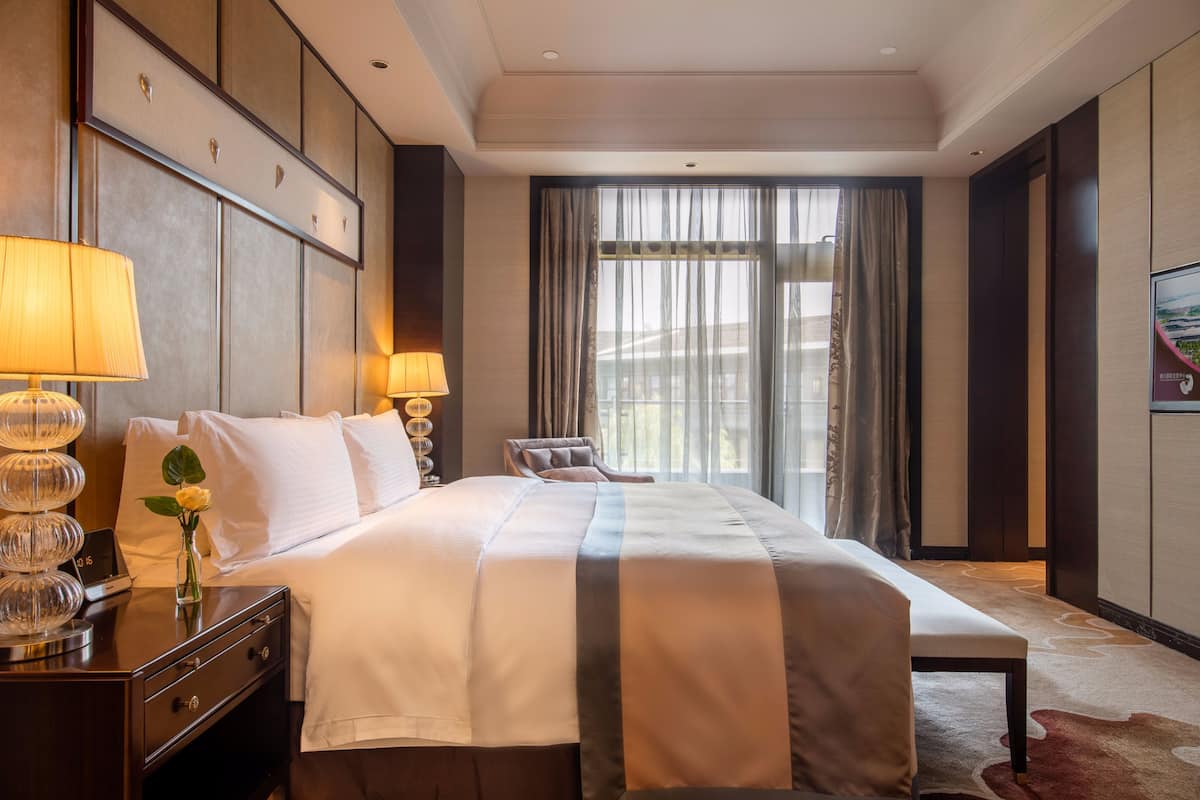 Ein Hotelzimmer mit einem großen Bett, Nachttischlampen, einem Beistelltisch, einem Sessel und Blick ins Freie durch das Fenster.