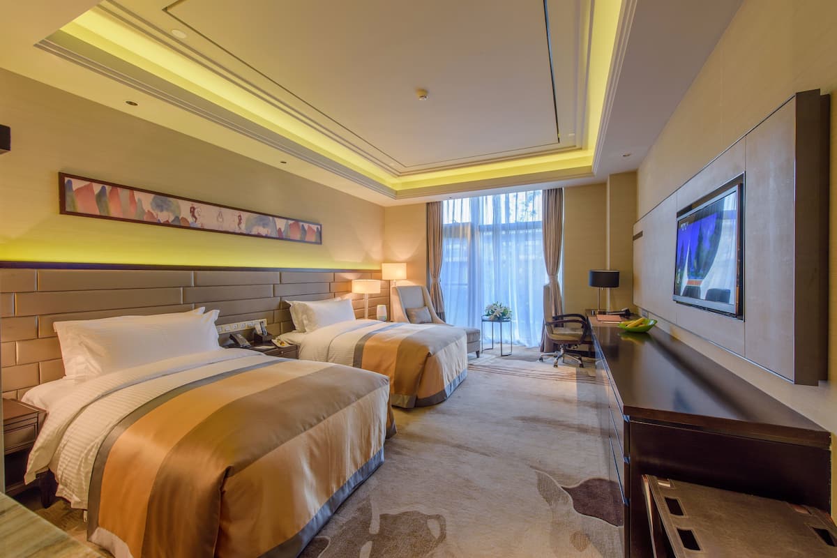 Ein Hotelzimmer mit zwei Betten, einem Schreibtisch, einem Fernseher und einem großen Fenster mit Vorhängen.