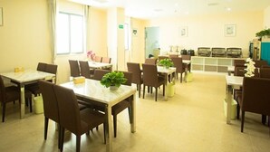 A dining area with multiple tables and chairs, a kitchen area in the background, and a window providing natural light. - GreenTree Inn SuZhou International Film City South JinShan Road Express Hotel (Suzhou)