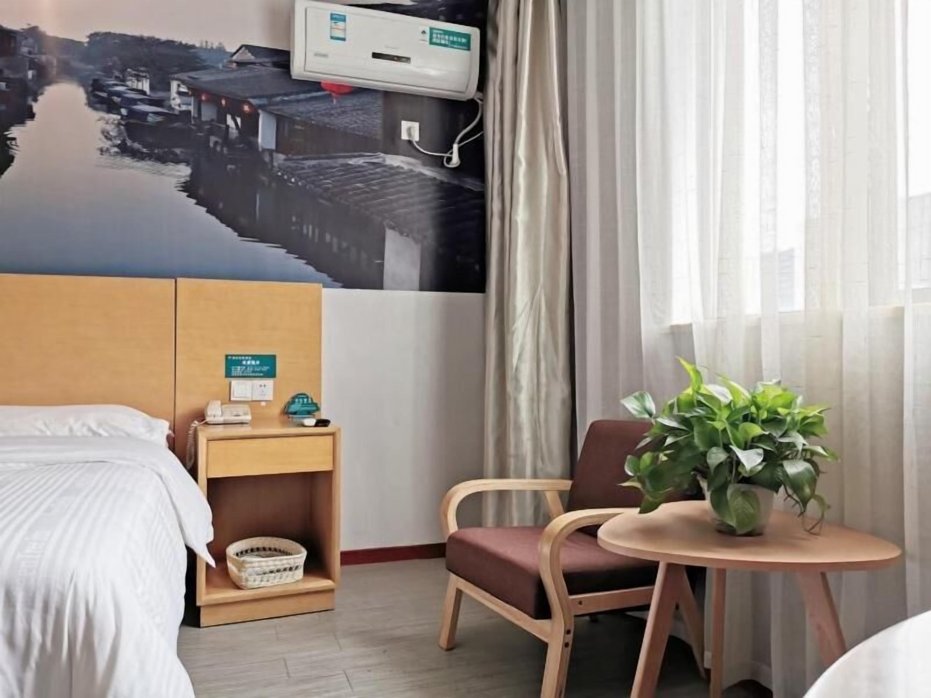 Une chambre d’hôtel comprenant un lit, un bureau avec un téléphone et une horloge, une chaise, une petite table avec une plante et un climatiseur fixé au mur.