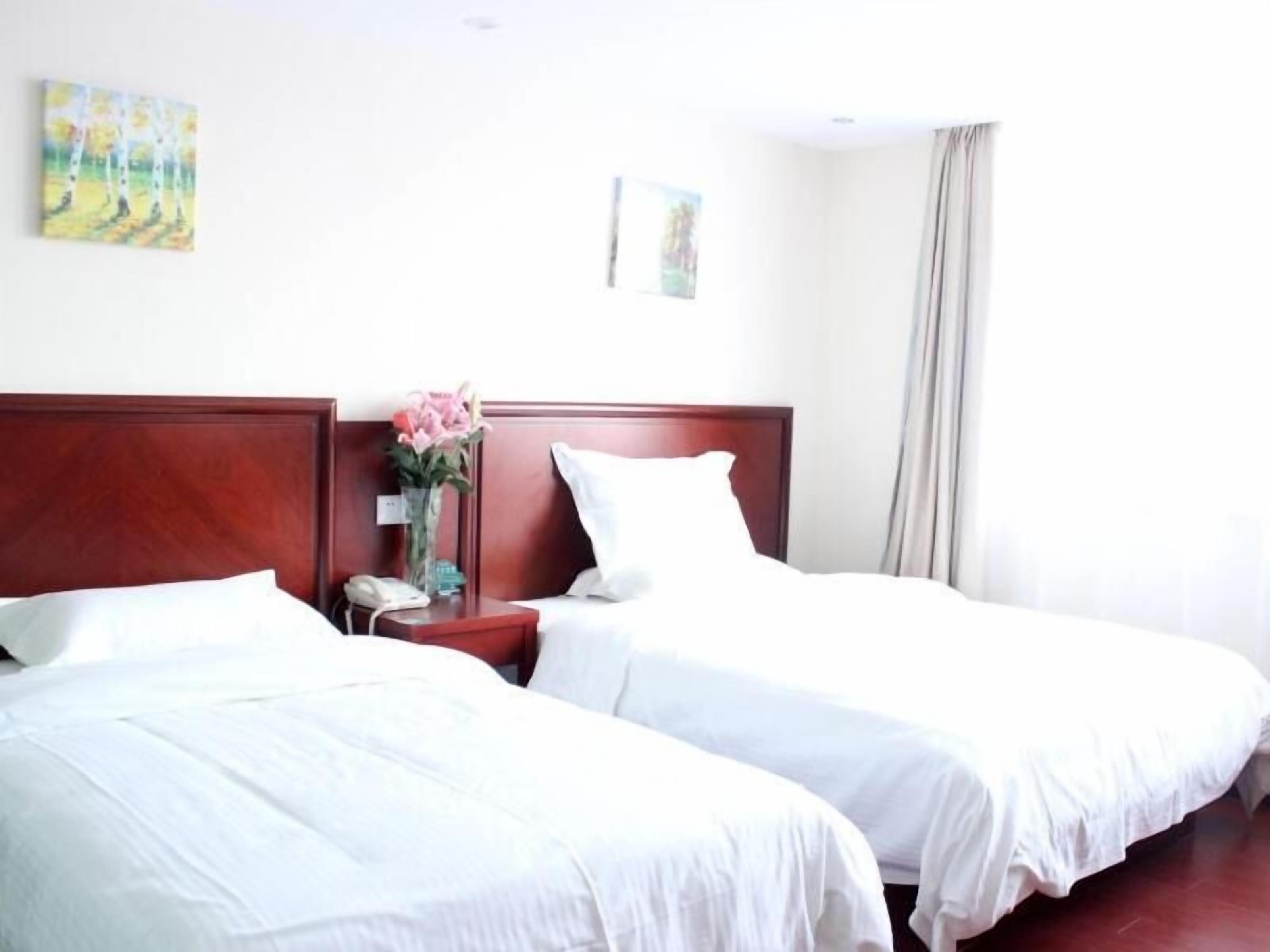 A hotel room with two single beds, a wooden headboard, a bedside table with a vase of flowers, and a painting on the wall.