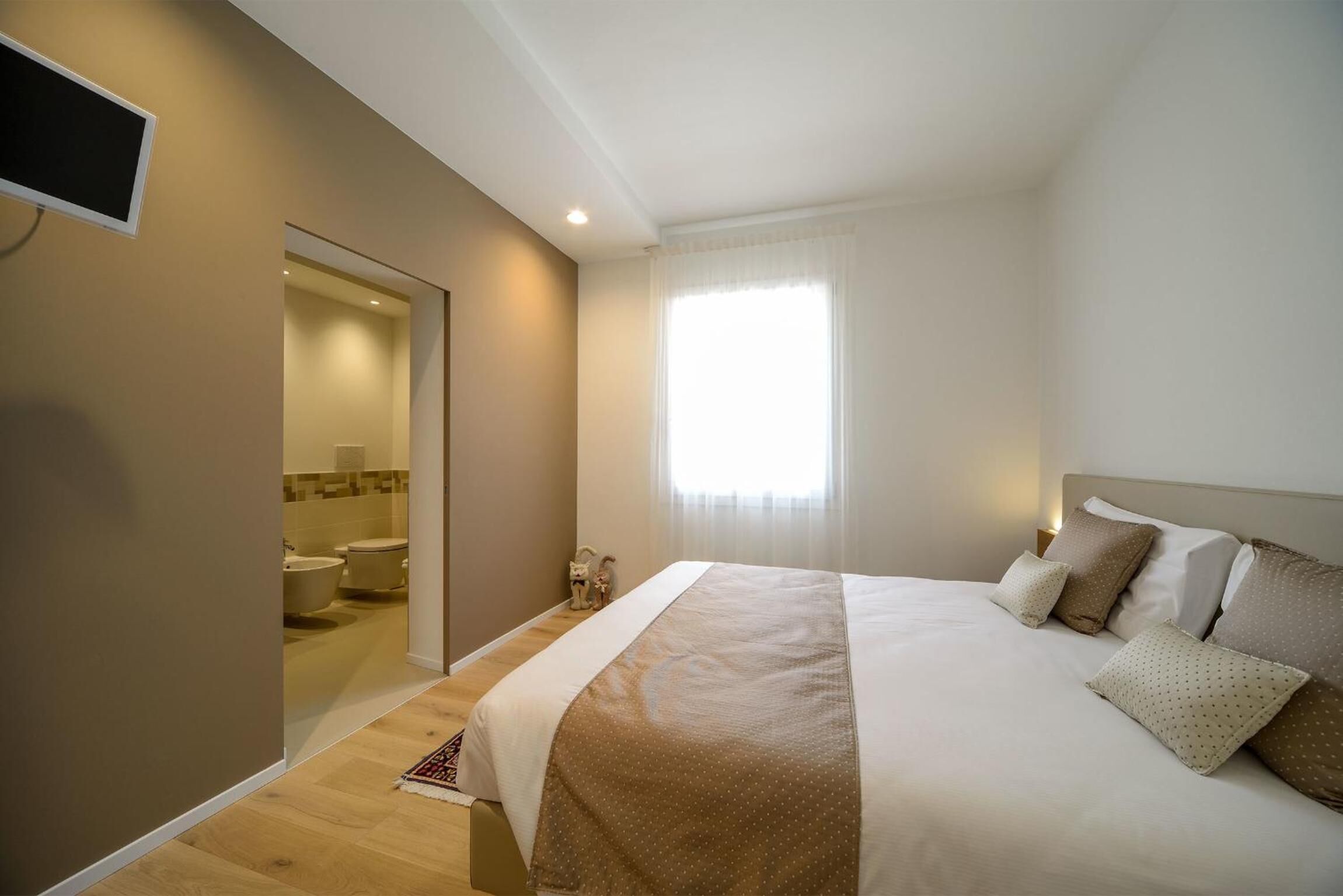 Ein Schlafzimmer mit einem großen Bett, einem an der Wand montierten Fernseher und einem durchgehenden Türrahmen einsehbaren Badezimmer.