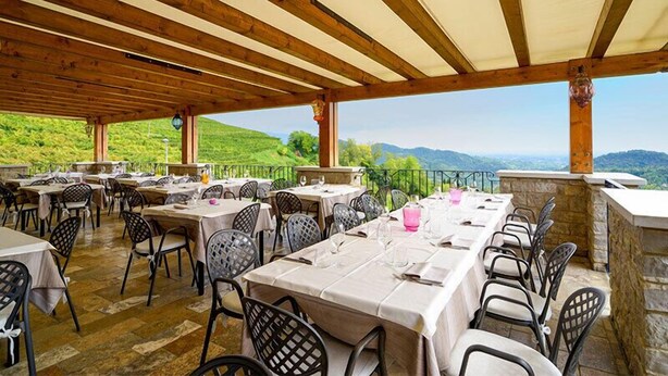 Un área de comedor al aire libre con mesas y sillas, techo de madera y vista a colinas verdes.