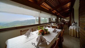Un comedor con mesas dispuestas para una comida, ventanas amplias con vistas a las montañas y vigas de madera en el techo.