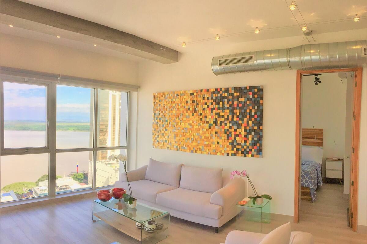 A modern living room with large windows, a glass coffee table, a white sofa, and a colorful abstract wall art piece.