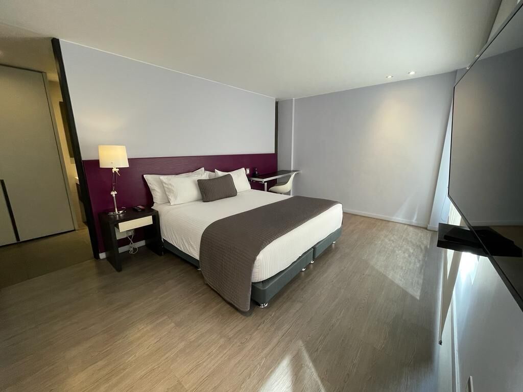 A modern hotel room with a large bed, a bedside table with a lamp, and a desk with a chair.