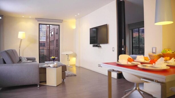 A modern living room with a sofa, a small table, a TV mounted on the wall, and a dining area with a red tablecloth. - Central Apartamentos Bogota (Bogotá)