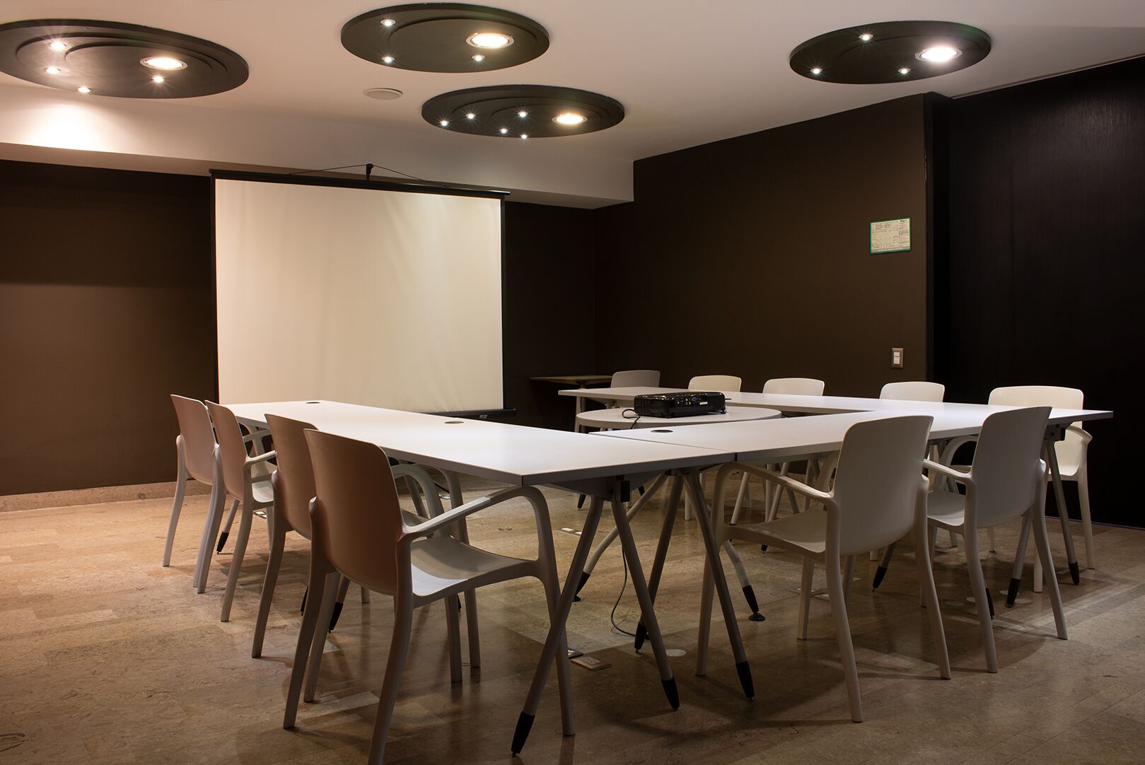 A conference room with a white table, white chairs, a projector screen, and recessed ceiling lights.