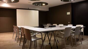 Une salle de conférence avec une table blanche, des chaises blanches, un écran de projection et des spots encastrés au plafond.