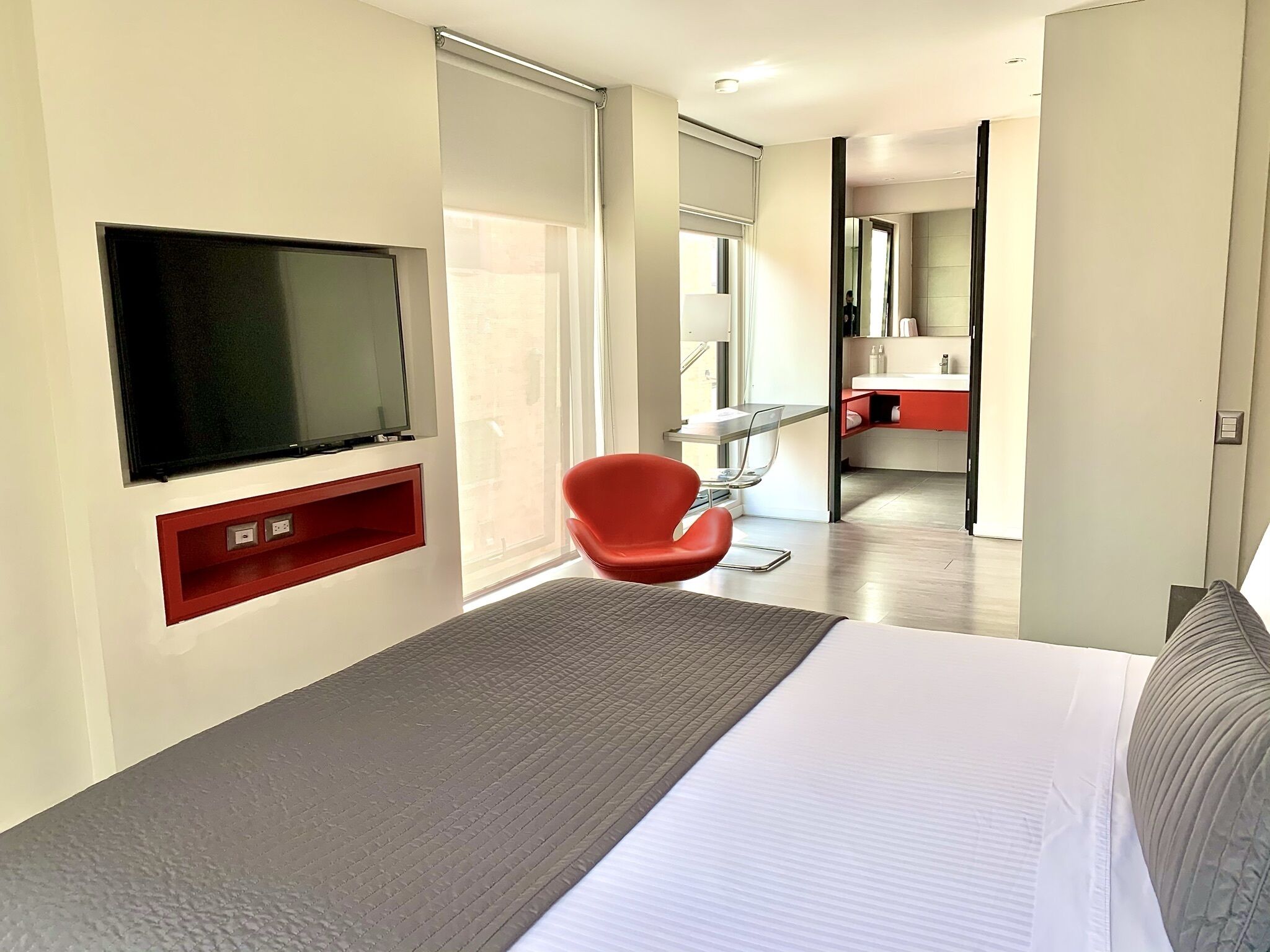 A modern hotel room with a large bed, a red chair, a TV mounted on the wall, and a bathroom visible through an open door.