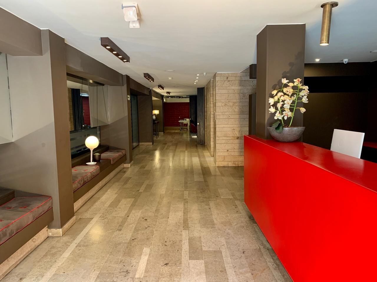 A hotel reception area with a red counter, a seating area with benches, and a hallway leading to a room with a red wall.