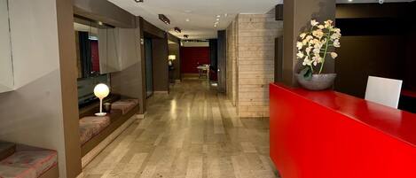 A hotel reception area with a red counter, a seating area with benches, and a hallway leading to a room with a red wall.