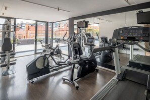A modern gym with various exercise equipment, large windows, and a view of buildings outside. - Central Apartamentos Bogota (Bogotá)