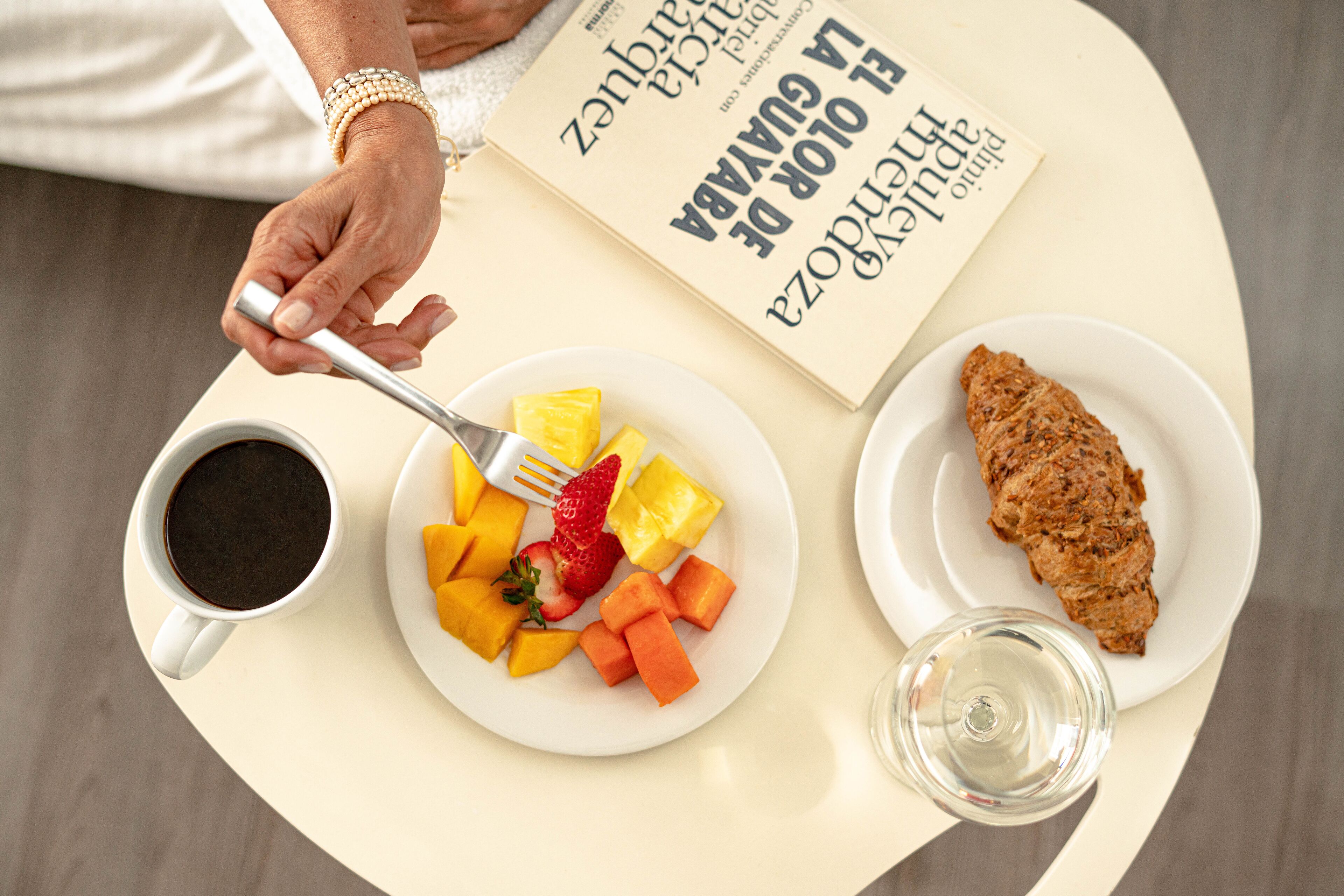 Een tafel met een schaal fruit, een kop koffie, een glas water en een boek.