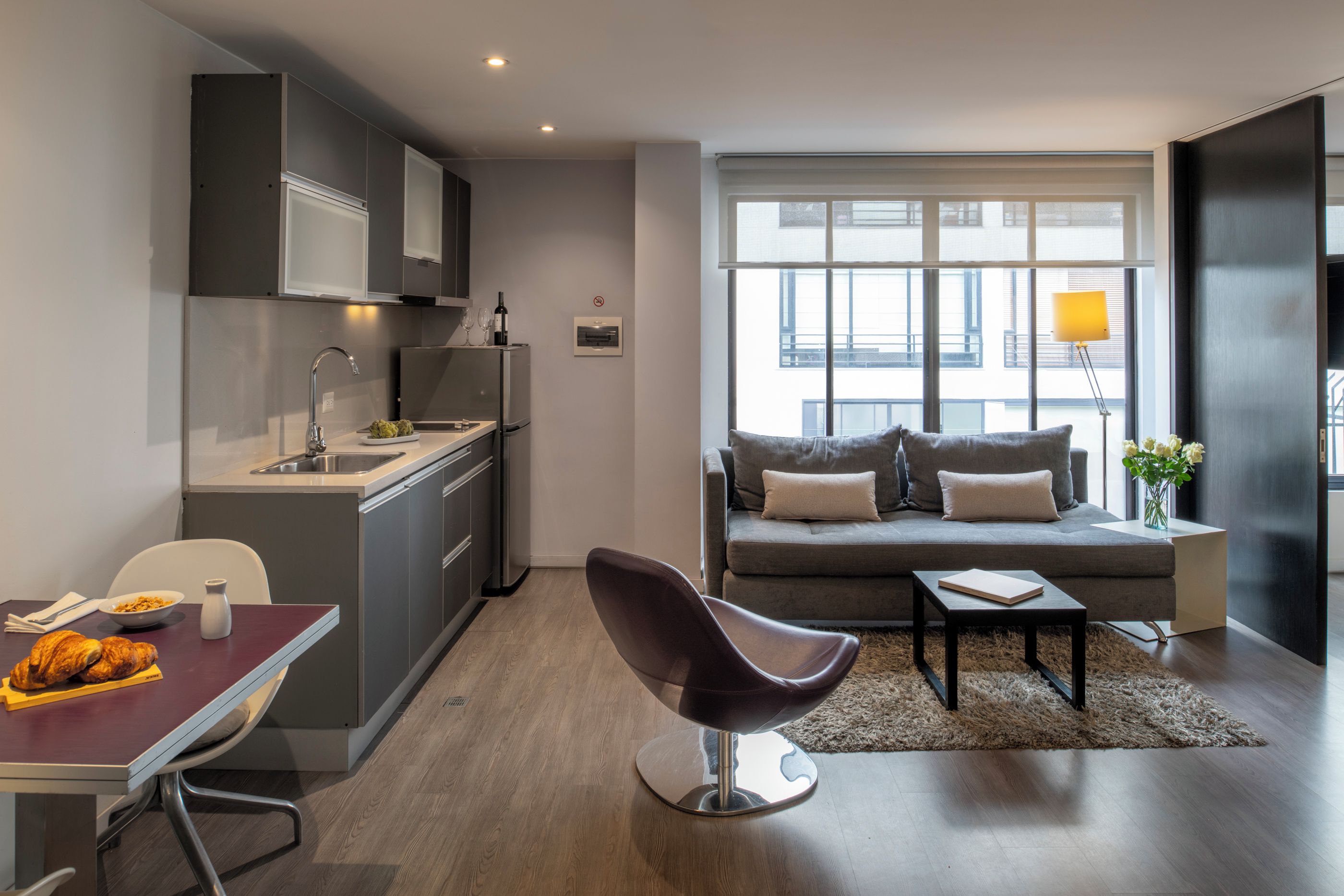 A modern kitchen with a dining area, a sofa with cushions, and a view of the city through large windows.