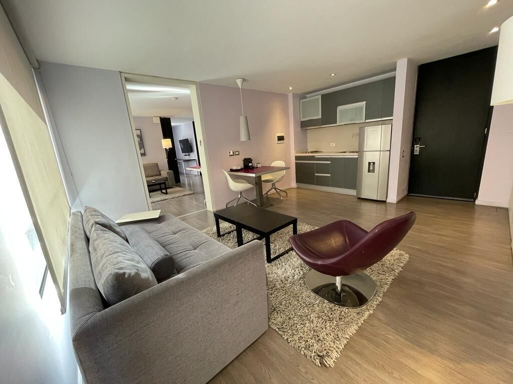 Un salon moderne avec un canapé gris, un fauteuil rouge, une petite table et un coin cuisine à l’arrière-plan.