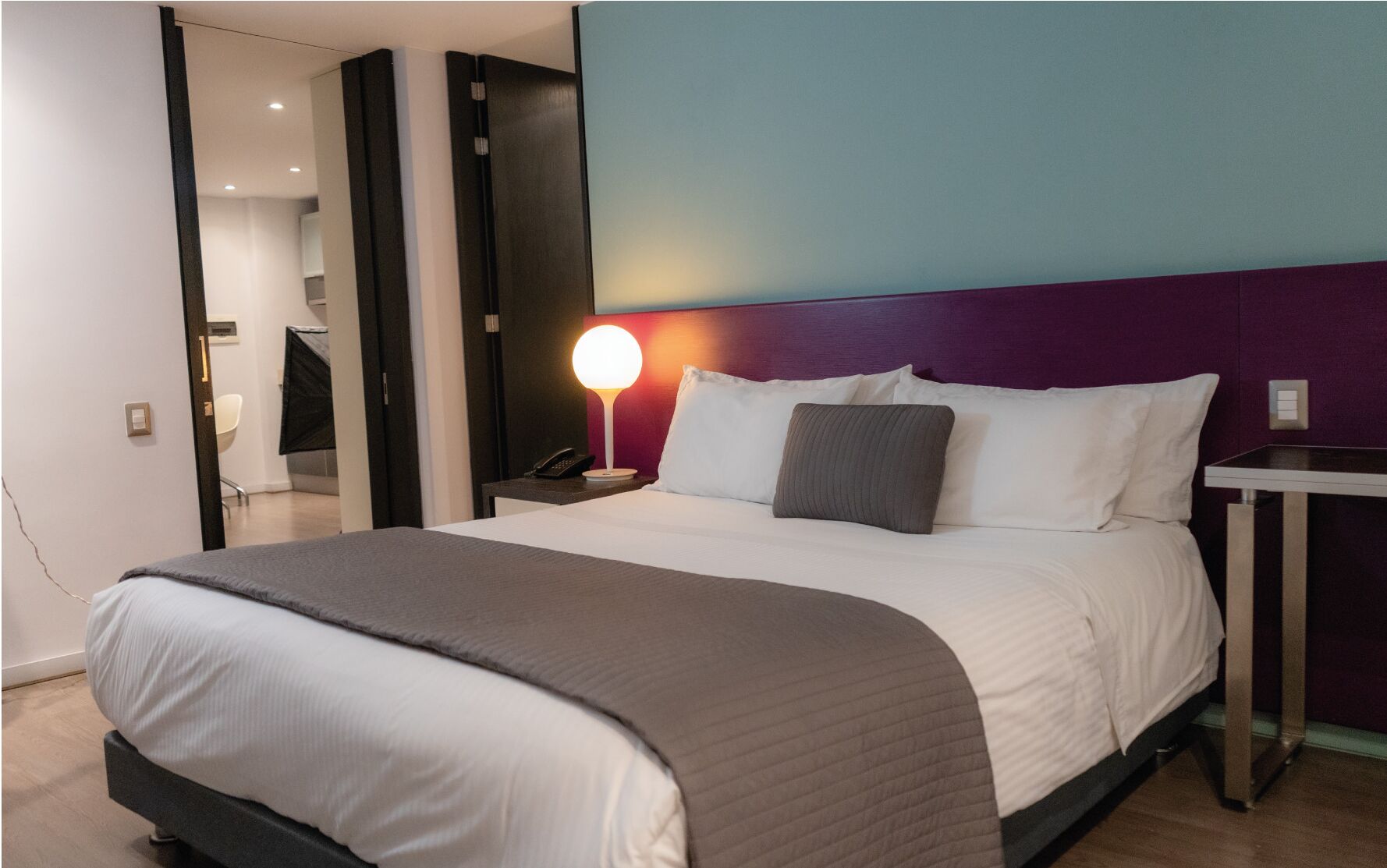 Ein modernes Hotelzimmer mit einem großen Bett, einem Nachttisch mit Lampe und Blick auf den Flur.