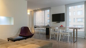 A modern living room with a sofa, a dining area with chairs, a TV mounted on the wall, and large windows offering a view of the city. - Central Apartamentos Bogota (Bogotá)