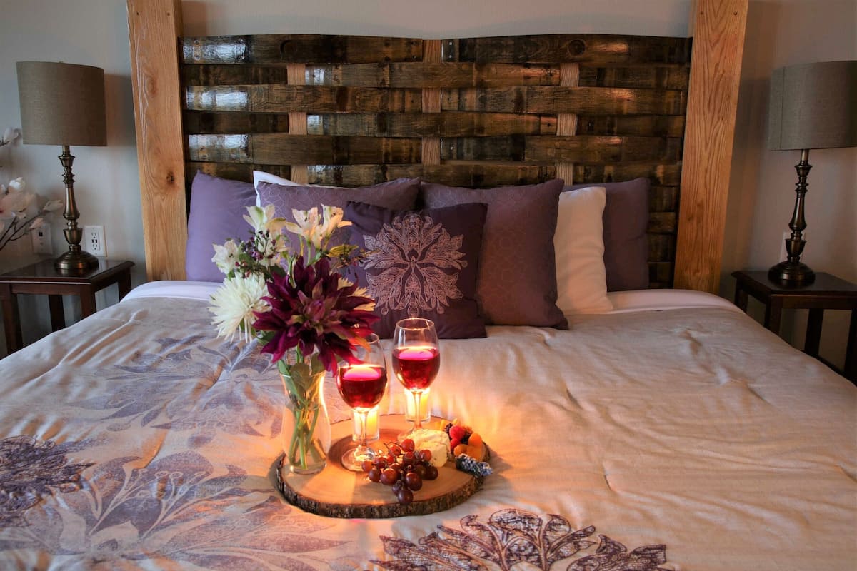 Een netjes opgemaakt bed met een decoratief dienblad met snacks en twee glazen rode wijn, een vaas met bloemen en een houten hoofdbord.