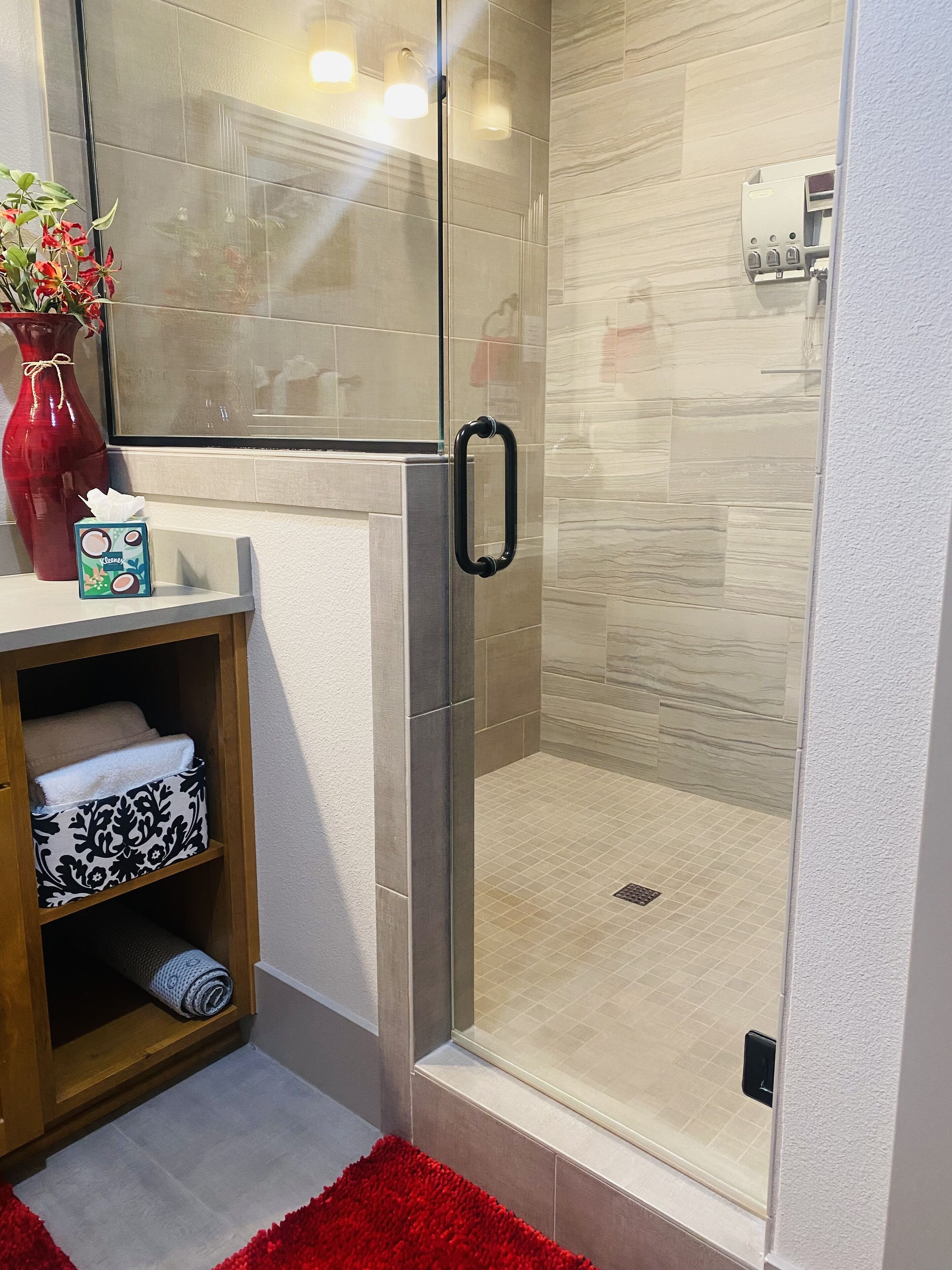 A modern bathroom with a glass shower enclosure, a wooden vanity with a vase and a tissue box, and a red rug.