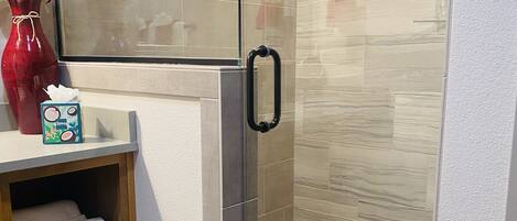 A modern bathroom with a glass shower enclosure, a wooden vanity with a vase and a tissue box, and a red rug.
