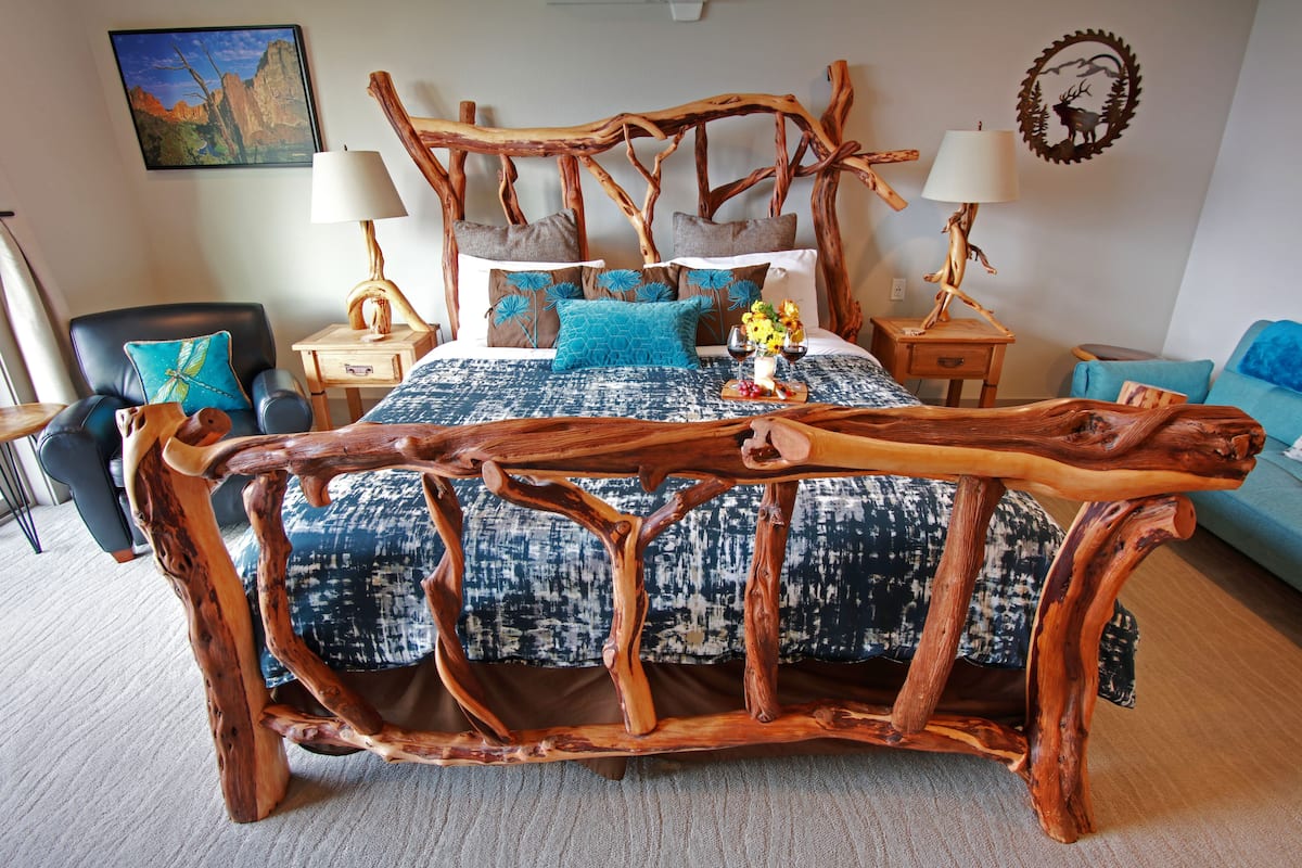 Een slaapkamer met een rustiek houten hoofdeinde, een bed met beddengoed met een patroon, twee nachtkastjes met lampen, een blauwe fauteuil en een aan de muur hangend schilderij.