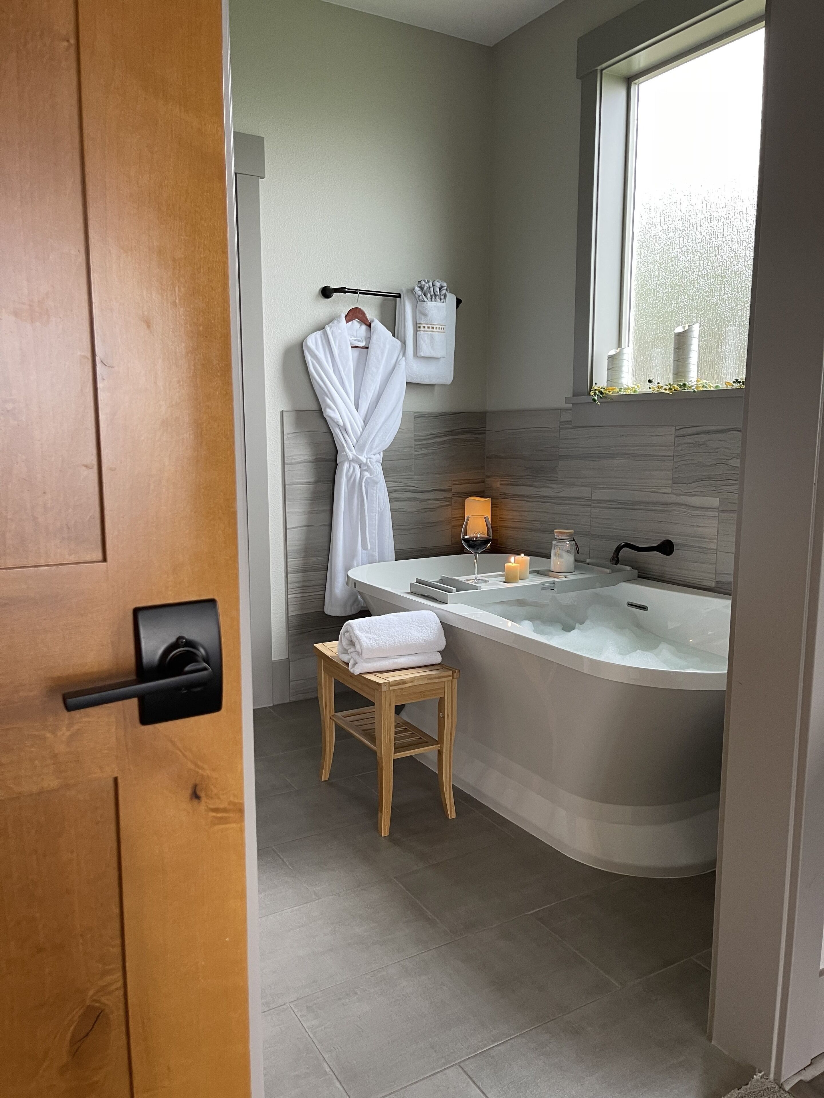 A modern bathroom with a freestanding bathtub, a wooden stool, a robe hanging on a towel rack, and a window with a view of the sky.