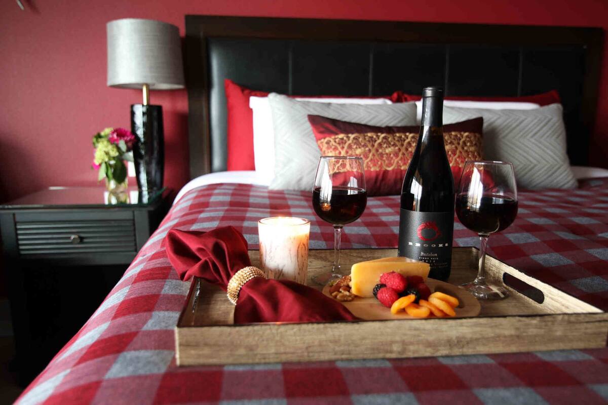 Een opgemaakt bed met een geruit beddekleed, een dienblad met kaas, fruit en wijnglazen, en een fles wijn op het bed.