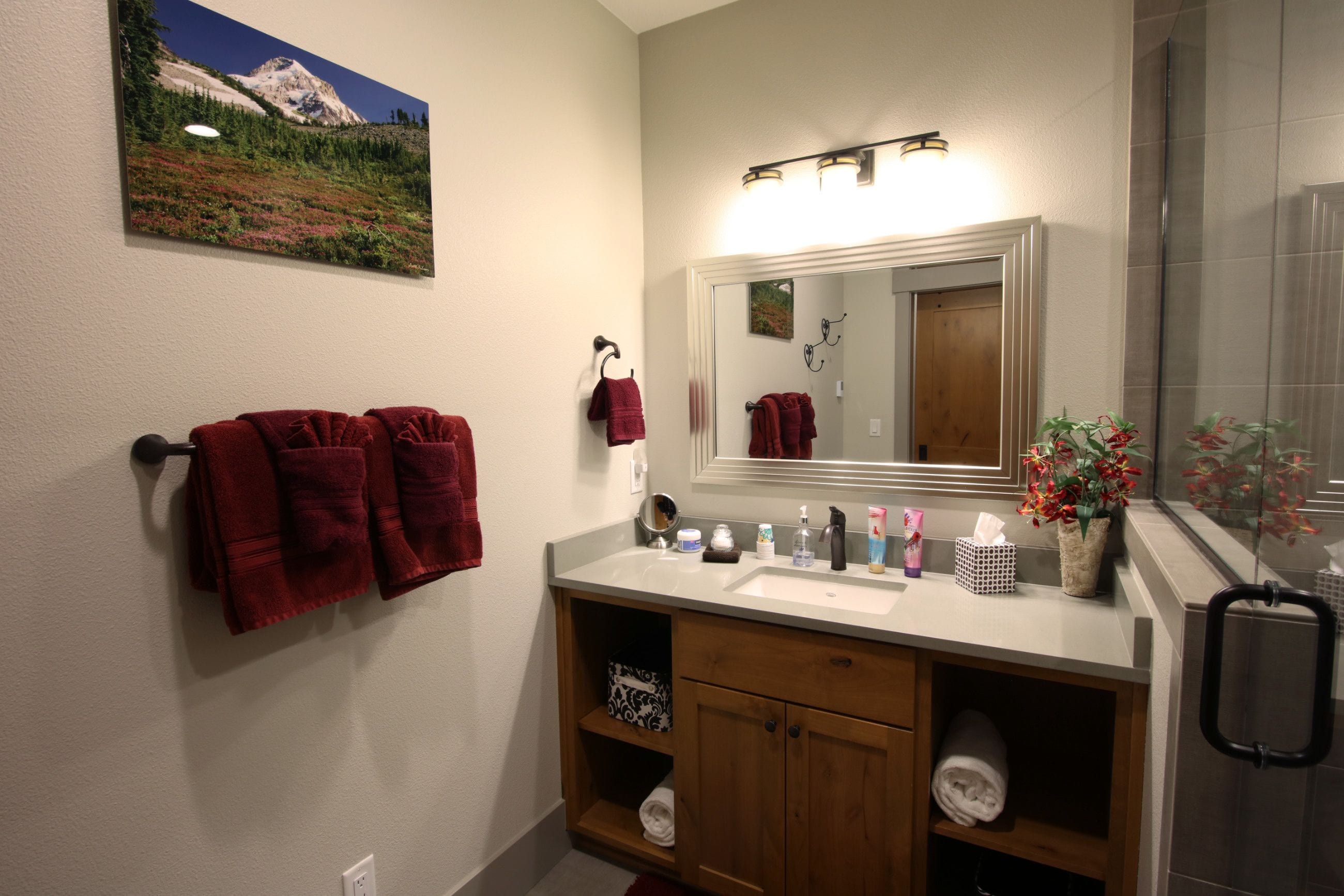 Une salle de bain avec un lavabo blanc, un meuble-lavabo en bois et un miroir au-dessus. Des serviettes rouges sont suspendues à des crochets et un tableau encadré est accroché au mur.