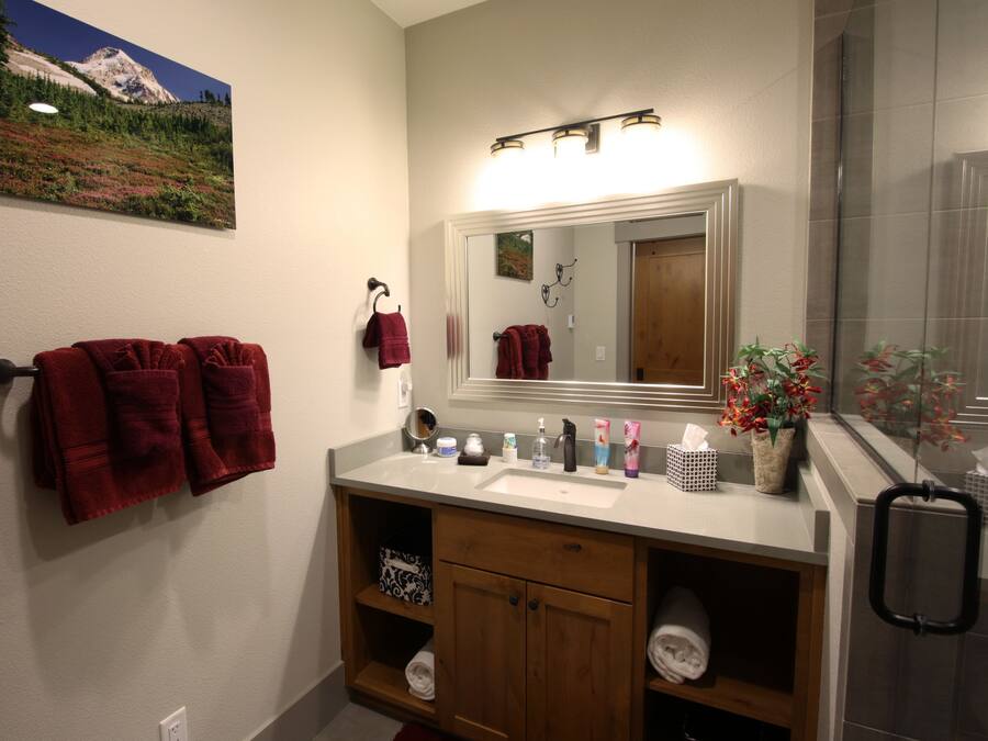 Une salle de bain avec un lavabo blanc, un meuble-lavabo en bois et un miroir au-dessus. Des serviettes rouges sont suspendues à des crochets et un tableau encadré est accroché au mur.