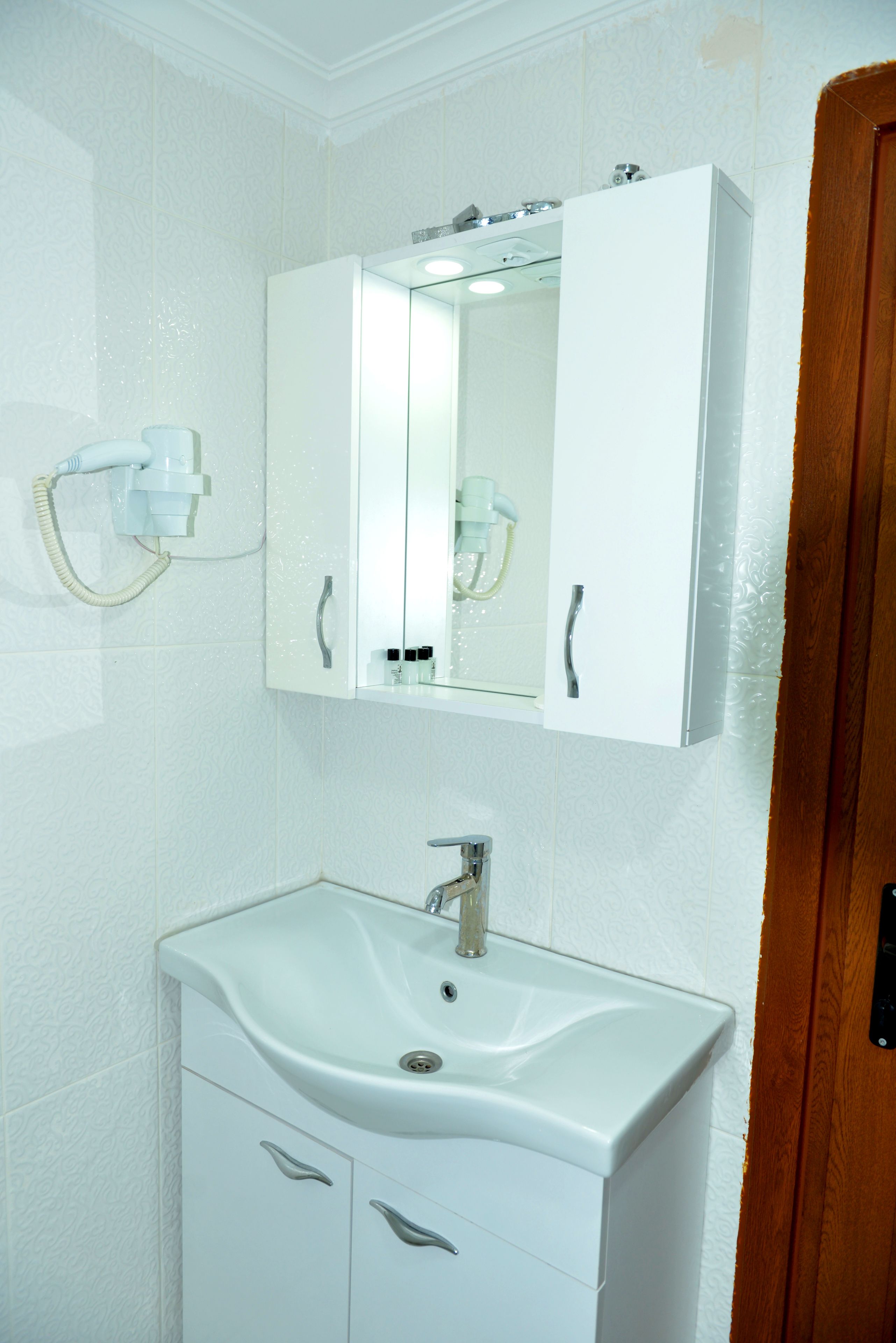 Un baño con lavamanos blanco, espejo y secador de pelo montado en la pared.