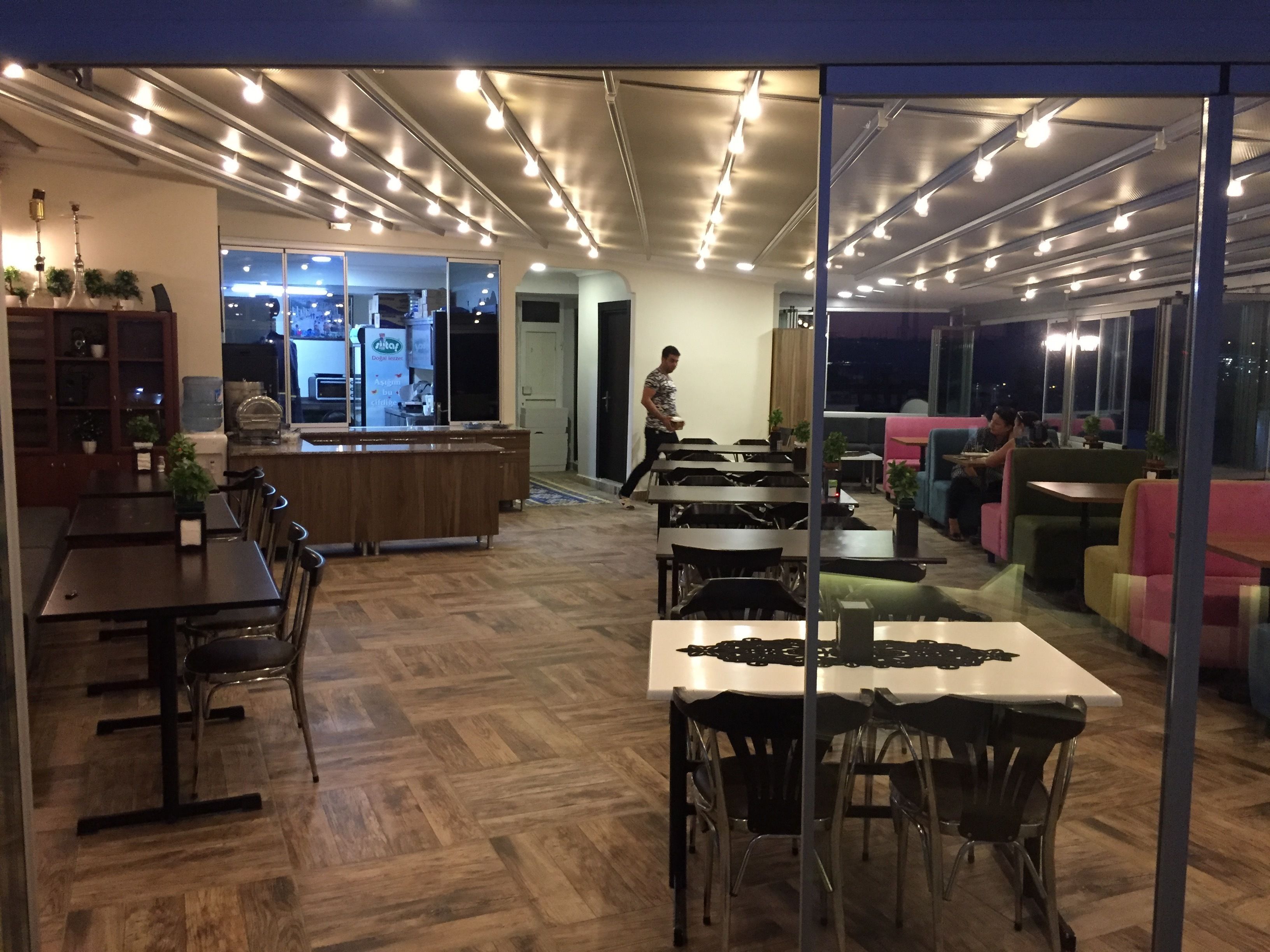 A modern dining area with wooden flooring, tables, and chairs. There is a counter with a menu and a person serving. The room has large windows and a view of the city at night.