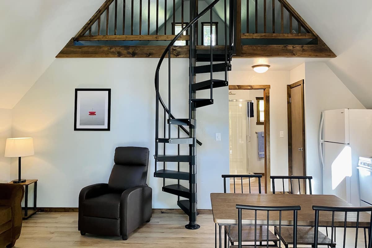 A loft-style room with a wooden staircase, a dining area with a table and chairs, a black leather chair, and a framed picture on the wall.