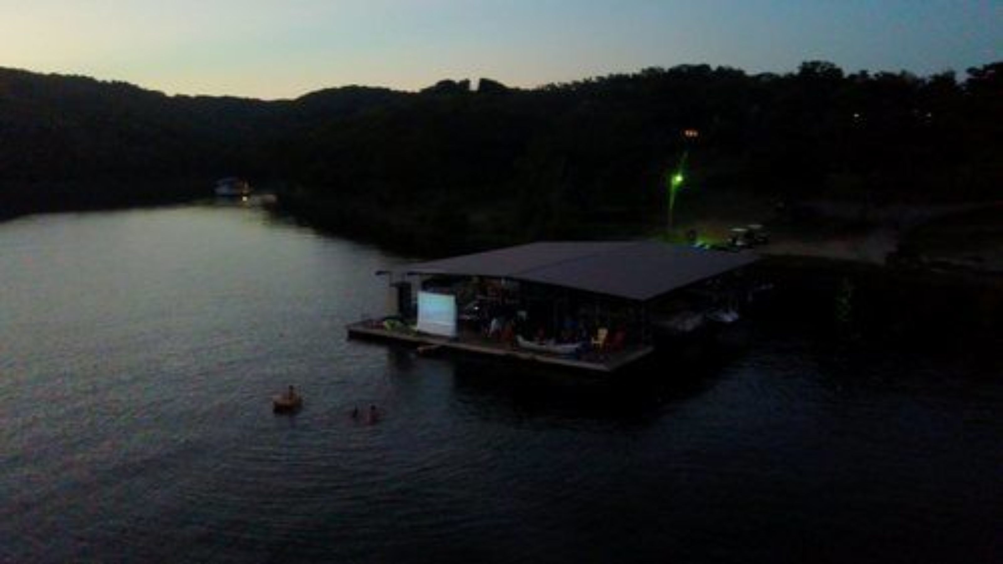 A docked boat with a covered structure, calm water, and a forested hillside in the background.