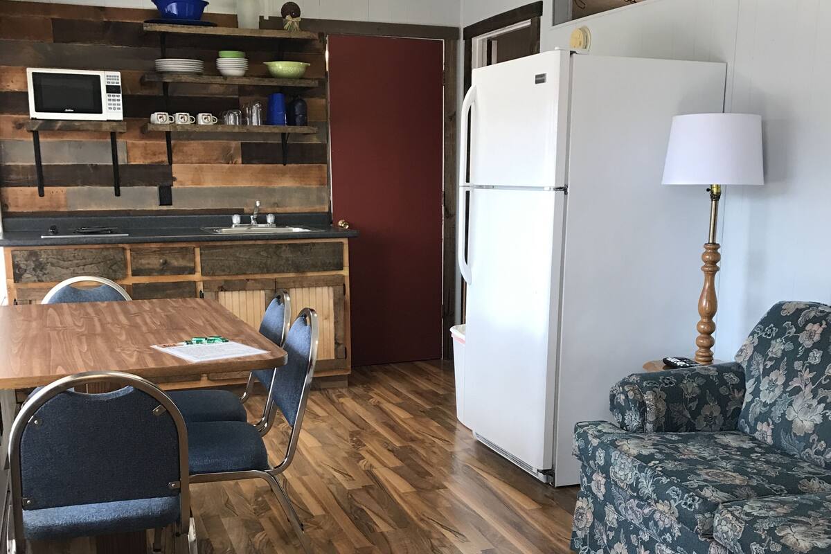 A compact kitchen with a microwave, sink, and refrigerator. There is a dining table with chairs and a floral-patterned sofa.