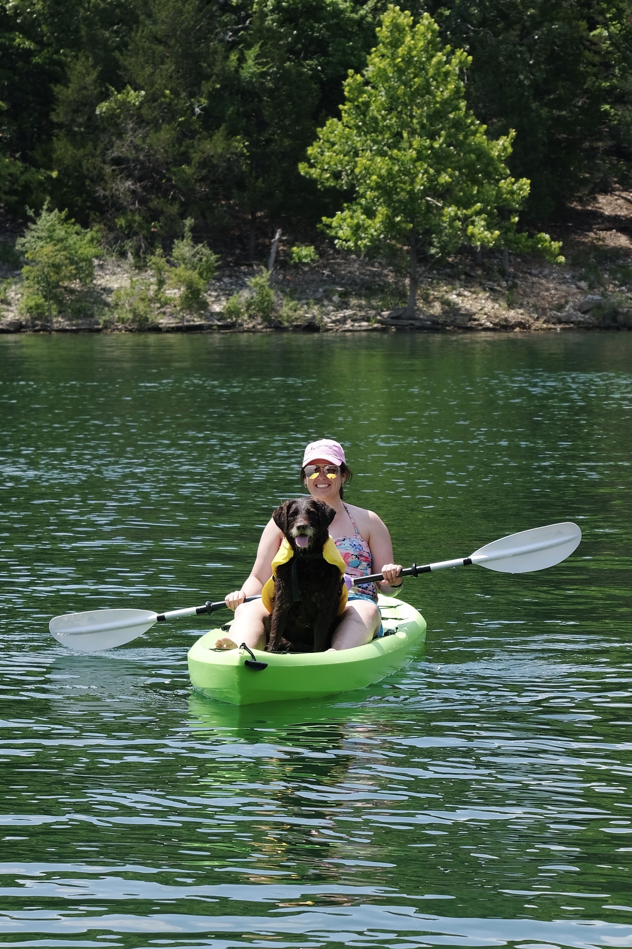 A person in a kayak with a dog, surrounded by calm water and greenery.