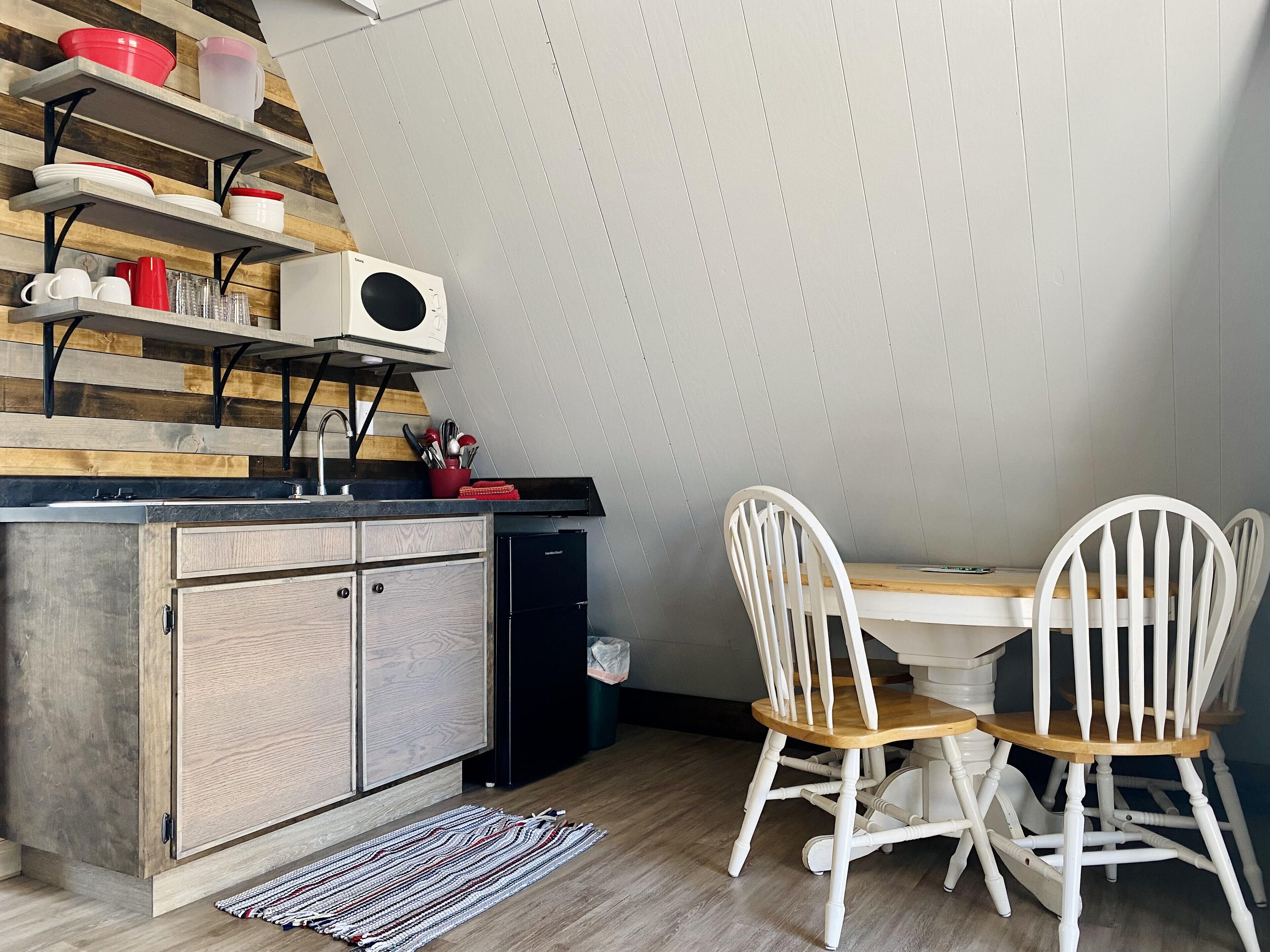 Una cocina compacta con microondas integrado, estantes de madera y una zona de comedor con sillas blancas.