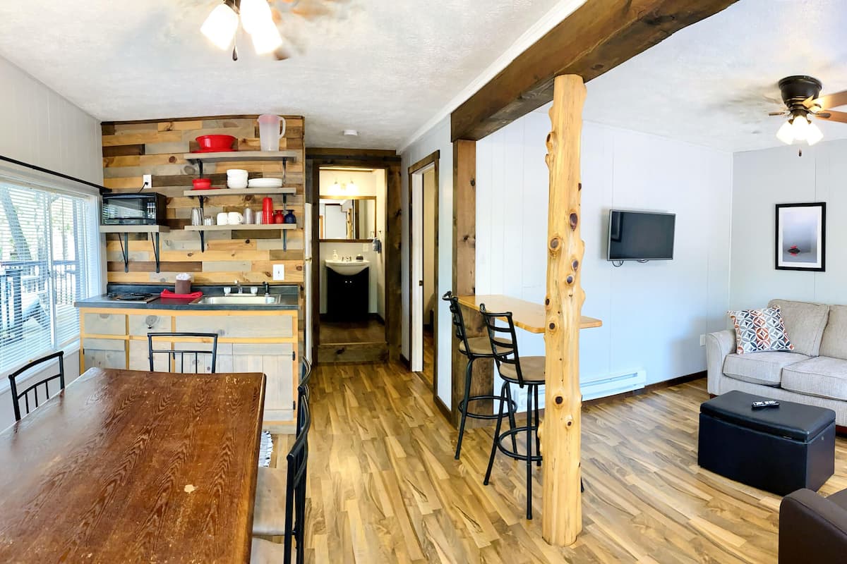 A modern kitchen with wooden accents, a dining area with a wooden table, and a living room with a sofa and a ceiling fan.