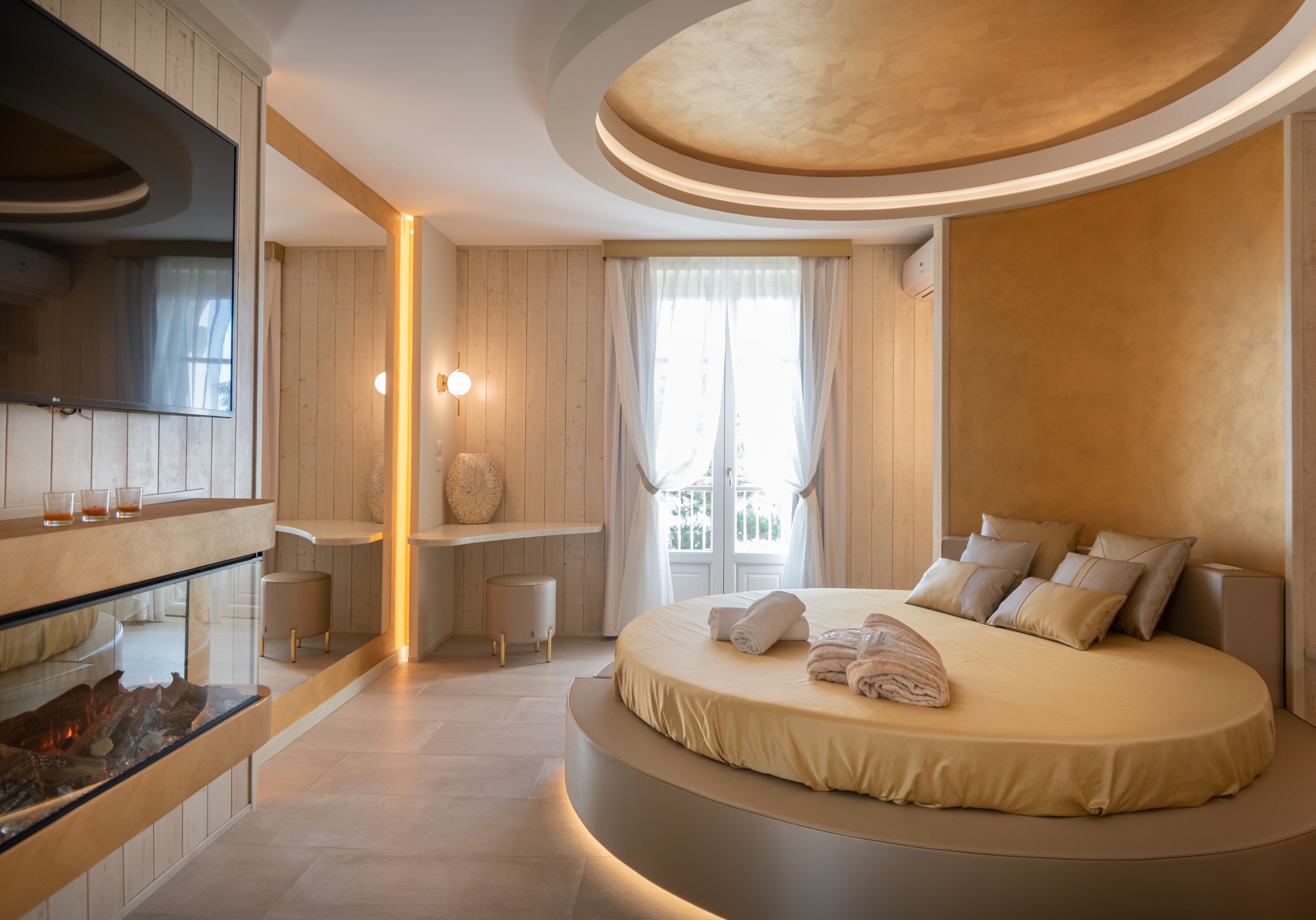 A modern bedroom with a large bed, a built-in TV, a vanity with a mirror, and a small seating area.