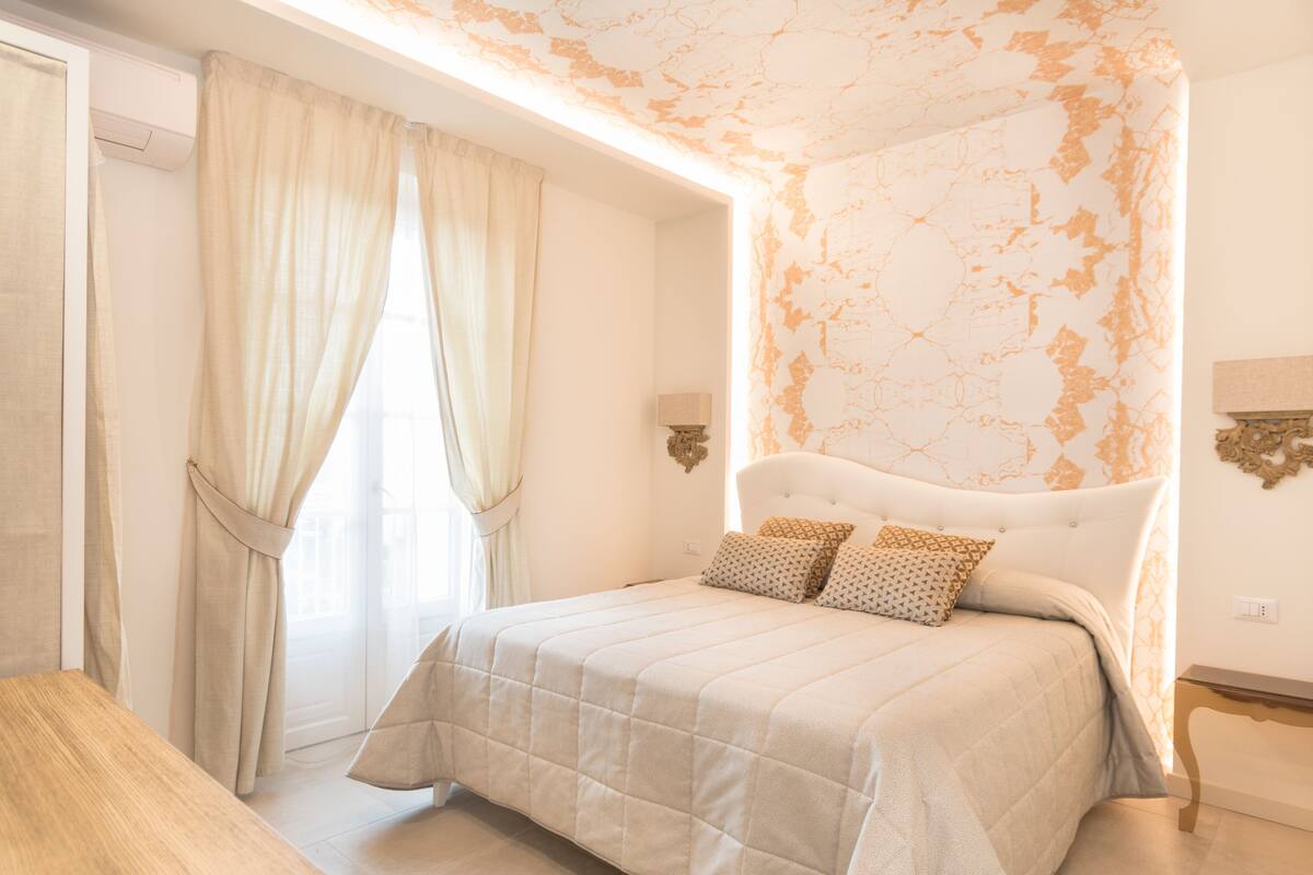 Quarto com cama, cortinas, mesa de cabeceira e luminária na parede.