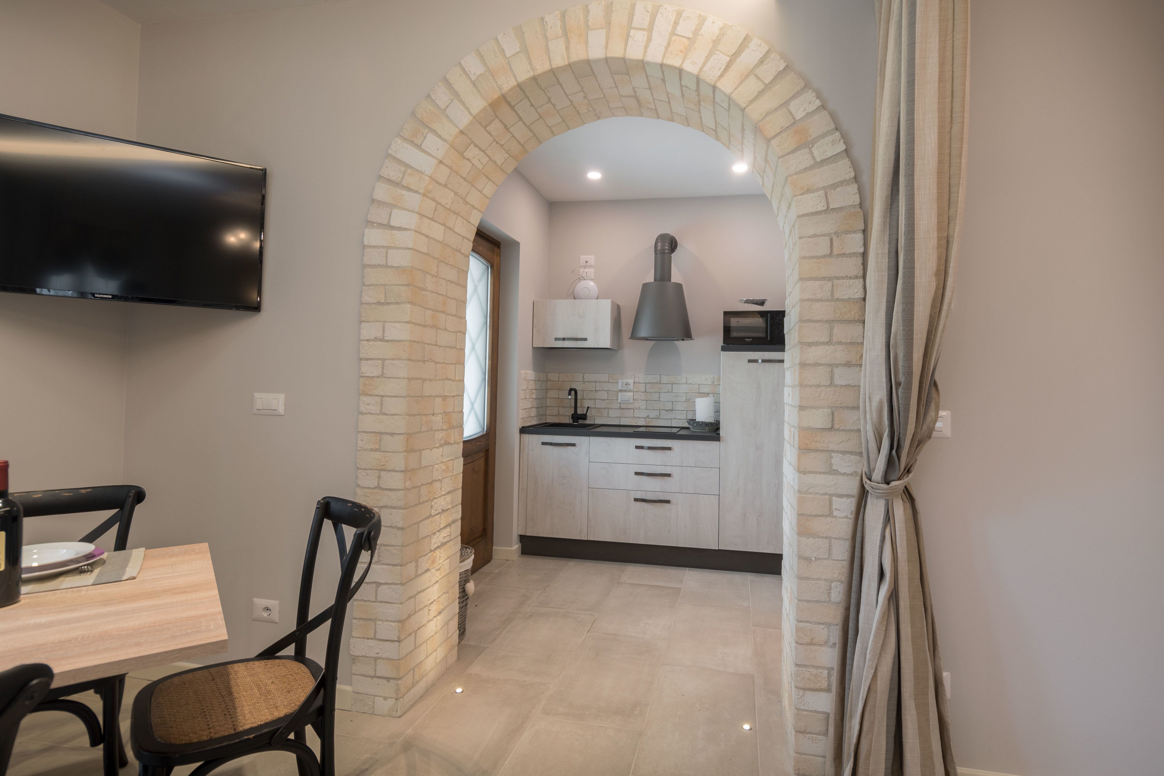 A modern kitchen with a brick archway, a flat-screen TV mounted above the counter, and a dining area with a table and chairs.
