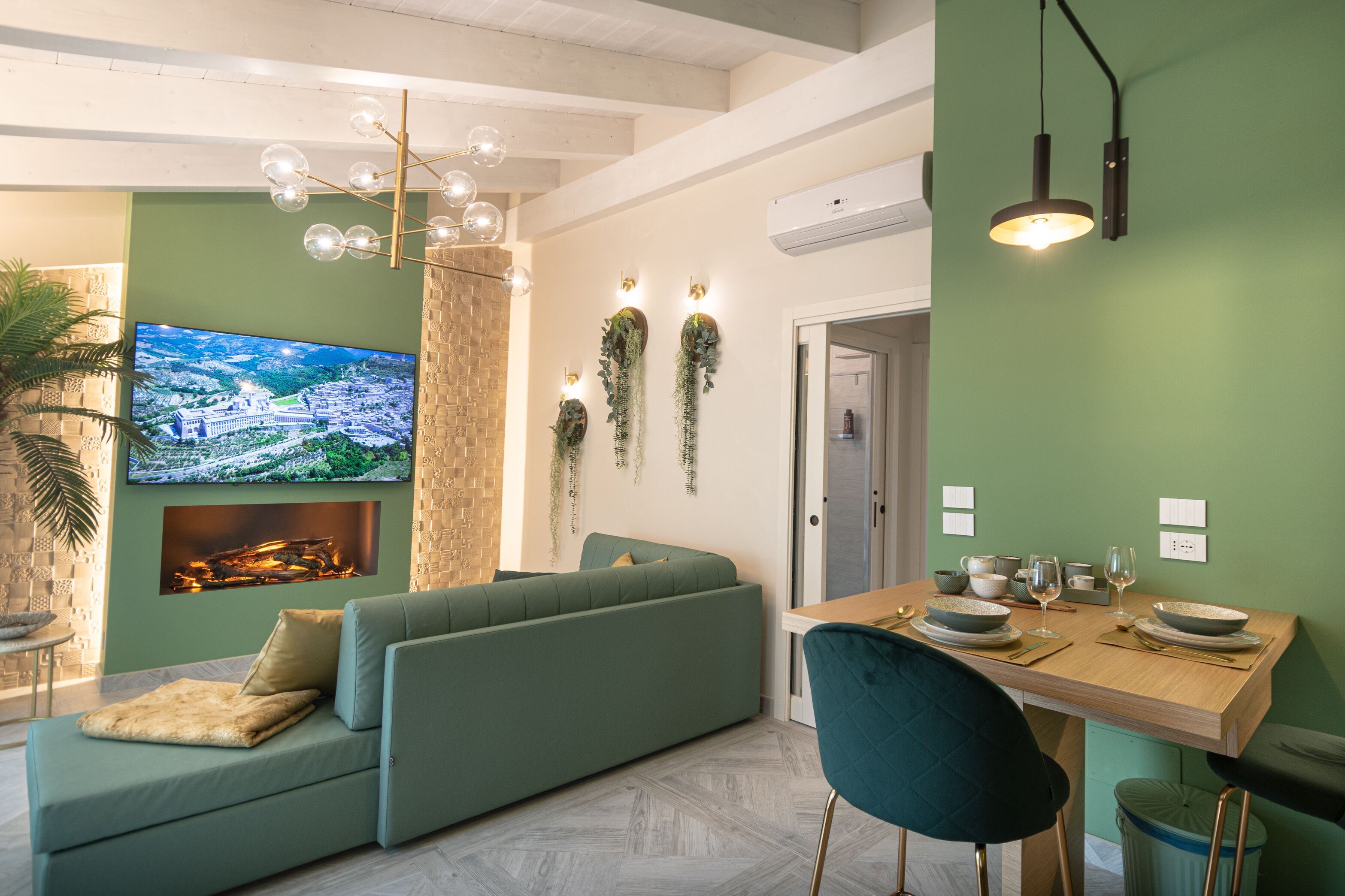 A modern living room with a green sofa, a dining table set for two, and a flat-screen TV mounted on the wall.