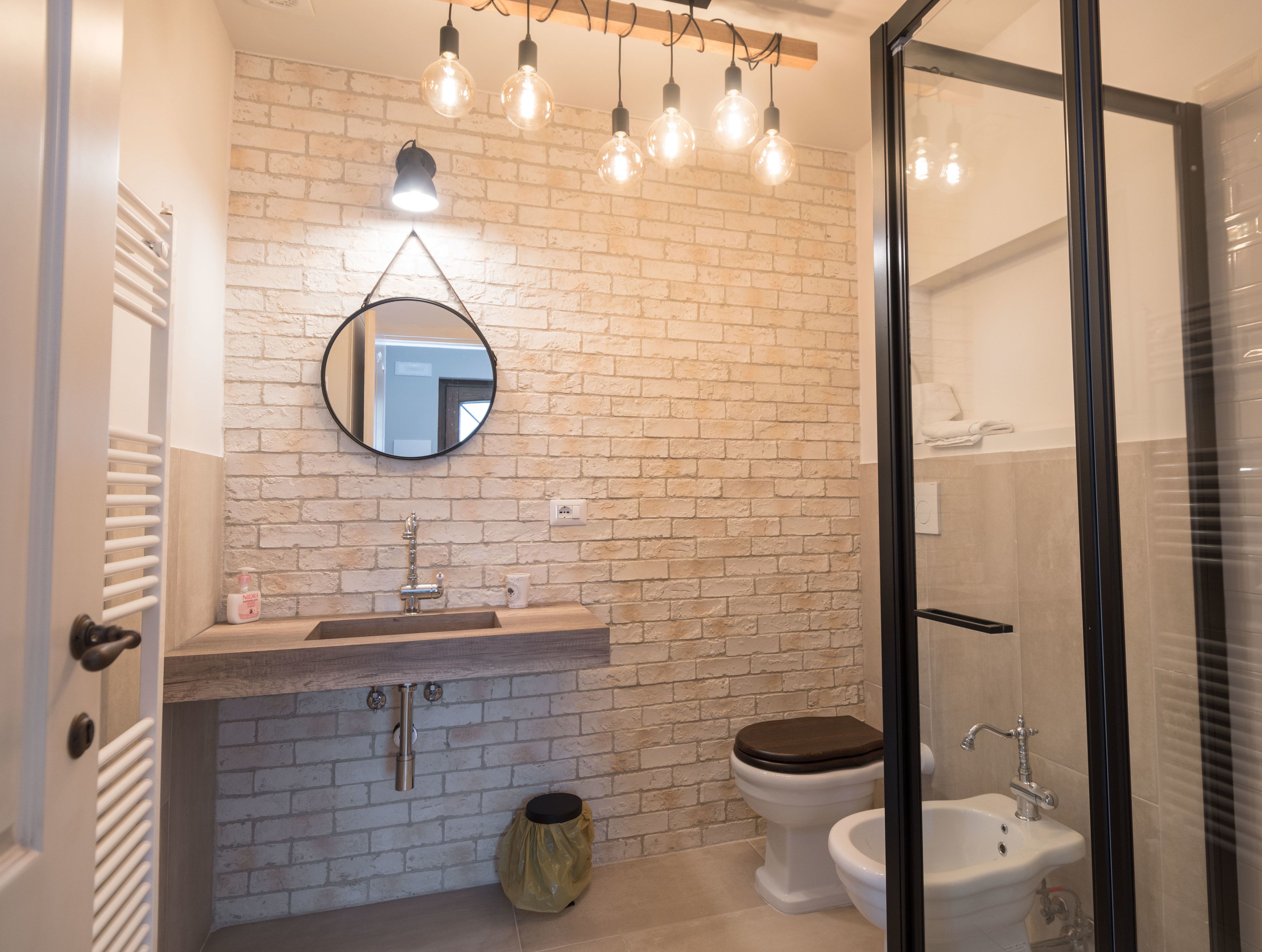 A bathroom with a white brick wall, a round mirror, a wooden vanity, a toilet, and a glass shower enclosure.