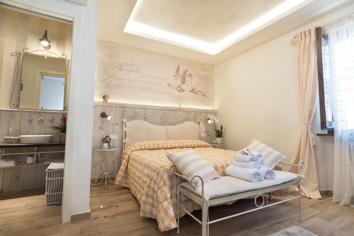 Quarto com cama, mesas de cabeceira, espelho, janela com cortinas e banheiro com pia e espelho.