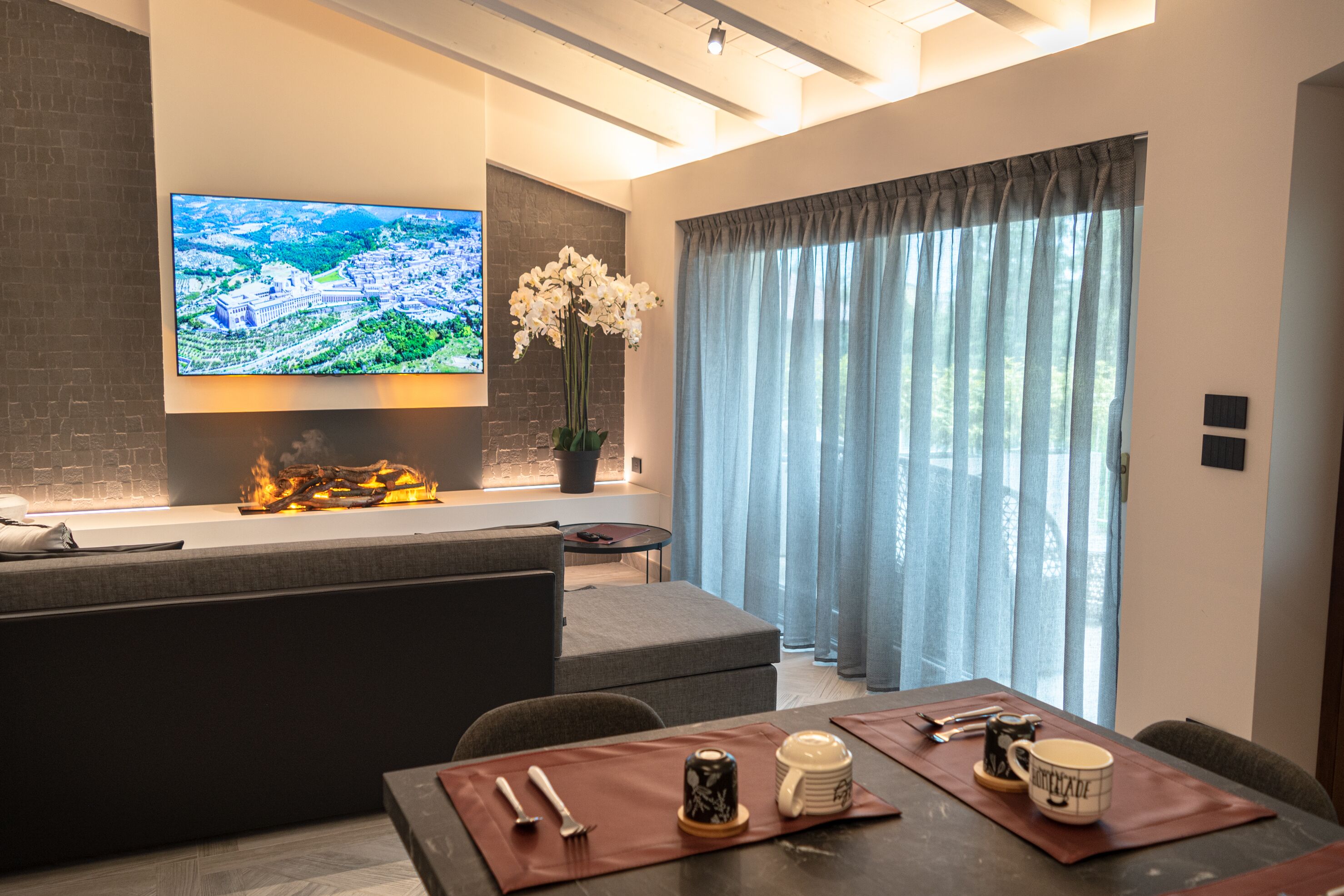 A modern living room with a flat-screen TV, a fireplace, a dining area with a table set for two, and a large window with curtains.